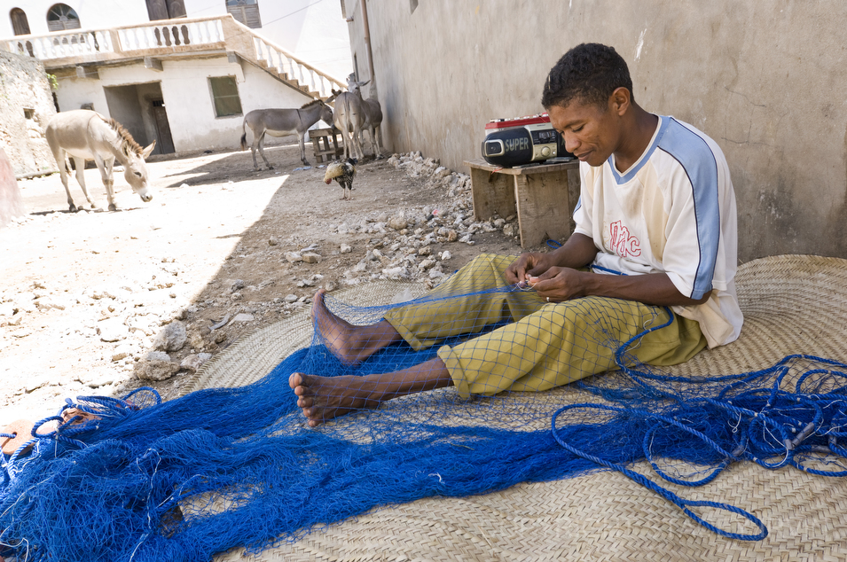 A fisherman repairs a net in Shela on Lamu Island while donkeys and chickens congregate in the background.