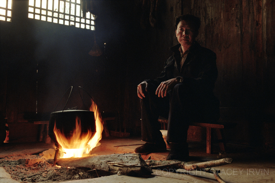 A man in Guanxi Province sits by the fire in his kitchen while heating water for tea.