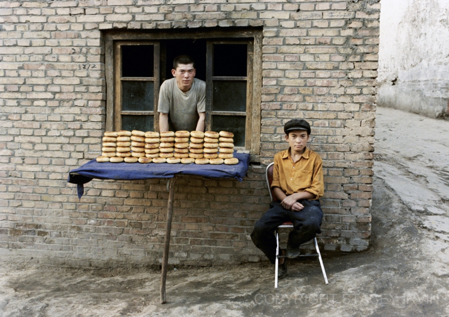 During my first trip to Kashgar I bought a bagel-like bread called girde nan from two handsome young men along a quiet lane. I photographed the bakers as I waited for their younger brother to fetch change for my purchase.

On my next visit in 2004, I brought this photograph to give to them. Unfortunately, their bread stand was vacant. An American man, who lives in Kashgar and speaks fluent Uighur, offered to help me search for the bakers. First we went back to the original location of the bakery, where the landlord told us that the older brother had moved back to his hometown outside of Kashgar. She thought that the younger brother still lived in town and told us to inquire at a nearby mosque, where we began what at first seemed to be a wild goose chase: first to the mosque, then to the Kenyi Bazaar, then the post office. We showed a photo around and eventually a woman recognized him. She pointed us around a corner and down a residential lane to a bread stand crowded with several old women. There he was, tucked inside a dark little room with cool white light filtering in from high above. He was perched on the top of an earth and brick oven platform. My heart was pounding as I handed him the photos! He recognized me and the photo, and his wonderful eyes were just the same. He nodded, set the photos down beside him as he worked, and smiled.
